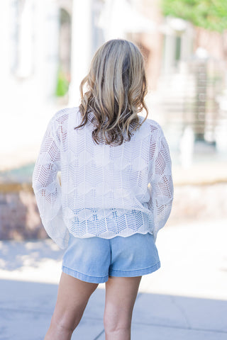 Back view of the Saltwater Breeze Sweater emphasizing the beautiful, continuous open-work chevron pointelle stitching. This angle highlights the relaxed, boxy silhouette, the airy movement of the long bell sleeves, and the playful scalloped hem resting just above the waistband of her light-wash denim shorts.