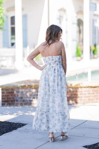 Back view of the Coastal Cloud Slit Dress showcasing the strapless design and premium, wide smocked elastic back panel that ensures a flexible, secure fit. The fluid chiffon skirt falls gracefully into a sweeping maxi length, finished with a romantic ruffled flounce hemline. The delicate watercolor-inspired floral print in muted periwinkle and soft sage is displayed beautifully as the model looks over her shoulder, styled casually with clear-strap wedge sandals.