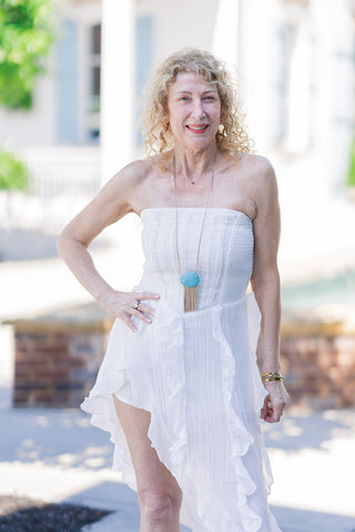 A portrait view of the Ibiza Breeze High-Low Dress focusing on the sleek, strapless tube bodice and straight-across neckline. The woman smiles warmly, posing with one hand on her hip to showcase the dress's flowy, lightweight fabric and the romantic vertical cascading ruffles that frame the daring one-side leg slit. She accessorizes the airy white dress with a striking turquoise stone pendant necklace featuring gold fringe.