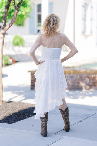 Back view of a woman wearing the pristine white Ibiza Breeze High-Low Dress. The image highlights the wide, comfortable back-smocked bodice that ensures a secure, form-fitting strapless fit. The lightweight woven skirt falls into a dramatic, asymmetrical high-low hemline finished with soft, cascading ruffle trim. She poses with her hands on her hips, styling the bohemian dress with brown western cowboy boots on a sunny outdoor walkway.