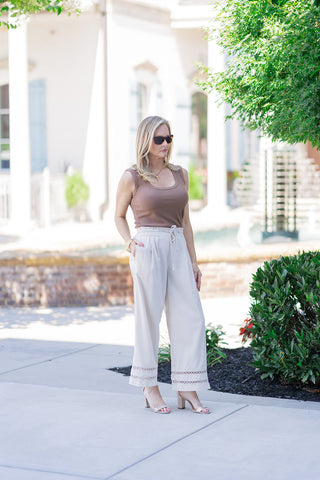 Woman posing outdoors in the Desert Linen Eyelet Pants, featuring a flowy, relaxed wide-leg silhouette in a light, natural linen blend. The pants are styled with a tucked-in earthy brown ribbed tank top, dark sunglasses, and metallic gold block-heeled sandals for an elevated coastal look. The high-rise elasticized ruffle waistband with an adjustable drawstring tie and the intricate geometric eyelet cutout panels along the lower hems are prominently displayed.