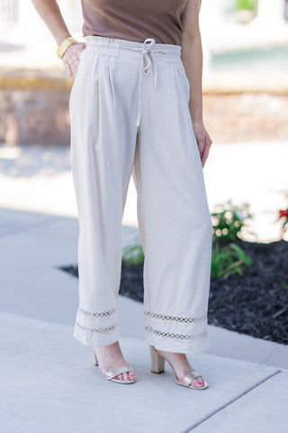 A closer front-angle view of the Desert Linen Eyelet Pants focusing on the garment's tailored details. The image highlights the high-rise, elasticized gathered ruffle waistband with a matching adjustable fabric drawstring and structured front pleats that enhance the wide-leg drape. The delicate, open-stitch geometric eyelet cutout trim encircling the lower hems adds a breezy, artisanal touch.