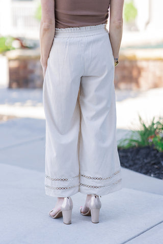 Back view of the Desert Linen Eyelet Pants showcasing the smooth, breathable drape of the premium lightweight linen-blend fabric. The stretchy, high-rise elasticized waistband ensures a comfortable, custom fit, while the wide-leg silhouette falls elegantly to the ankle. The airy geometric eyelet cutout detailing at the hemline provides a subtle hint of skin above metallic gold heels.
