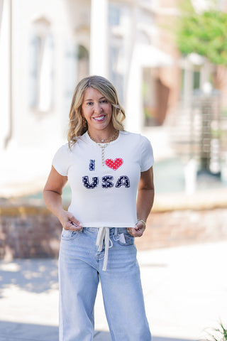 Front view of a woman posing casually in the All-American Girl Graphic Tee, a white baby tee with a comfortable ribbed crew neck. The shirt features a standout "I ❤️ USA" stitched applique graphic across the chest, with the letters crafted from navy blue and white polka dot fabric and the heart in vibrant red and white polka dots. Styled with layered gold necklaces and light-wash denim for a festive, patriotic summer look.