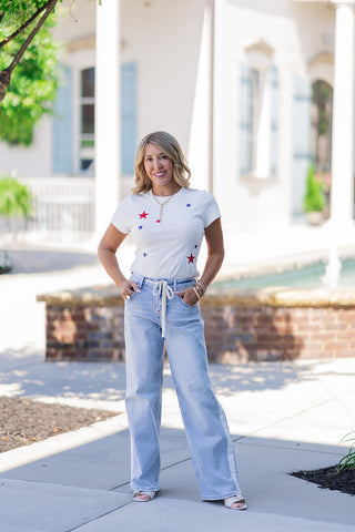 Woman posing outdoors in the Star-Spangled Firecracker Baby Tee, a crisp white short-sleeve top featuring playfully scattered, embroidered red and navy stars across the front. The modern baby tee is styled for a casual, festive summer look, tucked into high-waisted, light-wash wide-leg denim jeans with a white side stripe and drawstring waist. She is accessorized with layered gold necklaces and neutral slide sandals.