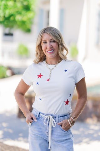 A portrait view of the Star-Spangled Firecracker Baby Tee highlighting the traditional ribbed crew neck and fitted short sleeves. The image captures the premium texture and visual depth of the beautifully stitched red and navy star embroidery against the clean white background. Styled with gold chain necklaces, the breathable cotton-blend fabric skims the body effortlessly without feeling restrictive.