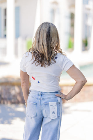 Back view of a woman wearing the Star-Spangled Firecracker Baby Tee, showcasing the flattering, slightly cropped fit that hits perfectly at the waist. The lightweight cotton-blend shirt features a few thoughtfully placed embroidered red and navy stars on the upper and lower back. It is paired with high-waisted, light-wash denim jeans, demonstrating a tailored length that pairs seamlessly without excess fabric or awkward bunching.