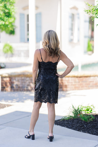Back view of a woman wearing the Noir Novelty Lace Dress, a black bodycon mini dress featuring delicate spaghetti straps and a clean, concealed center back zipper. The image highlights the intricate, heavy novelty lace overlay and the beautifully embroidered scalloped hemline. She is styling the elegant cocktail dress with black double-strap block heels, posing outdoors on a sunlit sidewalk with one hand on her hip.