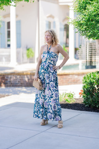 Full body front view of a woman smiling outdoors in the Lavender Lagoon Halter Jumpsuit. This dramatic, floor-skimming wide-leg jumpsuit creates the sweeping illusion of a maxi dress, featuring a vibrant, large-scale botanical print of lavender and violet lilies with deep teal green leaves on a crisp creamy background. Styled for an elegant resort look, the outfit is paired with neutral block-heeled sandals, gold bracelets,