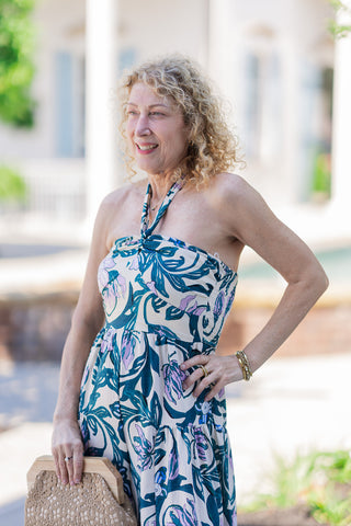 A closer portrait view of the Lavender Lagoon Halter Jumpsuit highlighting the sculpted sweetheart bust with a central twisted knot detail. The image showcases the supportive halter neck tie extending upwards and the premium lightweight woven fabric adorned with lush tropical florals. The model casually rests one hand on her hip while holding a textured woven clutch, perfectly capturing an effortless, bohemian glamour.