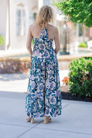 Back view of the Lavender Lagoon Halter Jumpsuit demonstrating the secure, customizable fit of the tied halter straps and the premium, wide smocked elastic back panel. The image captures the dynamic movement of the wide-leg trousers cascading into voluminous tiered ruffle panels, beautifully displaying the artistic botanical print from top to bottom. Styled with woven neutral block heels for a breezy summer aesthetic.
