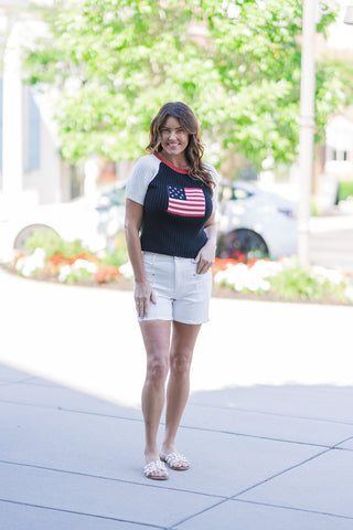 Woman modeling the Americana Heritage Ribbed Knit, a short-sleeve sweater featuring a deep navy blue vertically ribbed bodice with a central intarsia-knitted American flag emblem. The retro-inspired top is detailed with crisp white raglan sleeves and a vibrant red crew neckline. She is styling this patriotic knit with high-waisted white denim shorts and white slide sandals, posing outdoors on a sunny sidewalk.