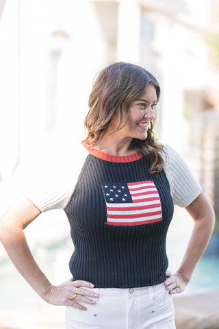 A portrait view of the Americana Heritage Ribbed Knit top, highlighting the textured detail of the central knitted American flag graphic with its stars and stripes. The woman poses with her hands on her hips, showcasing the form-fitting stretch of the vertical navy ribbing, the sporty white raglan short sleeves, and the contrasting red neckline.