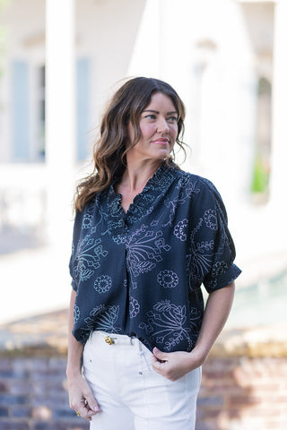 A portrait view of the Line-Art Lily Blouse highlighting the elegant split v-neckline framed by an intricately structured ruffled collar. The image showcases the top's fine slub texture and the relaxed drape of the dolman sleeves. The classic black-and-white botanical doodle print provides a polished, high-end boutique aesthetic as the blouse is partially tucked into white denim.