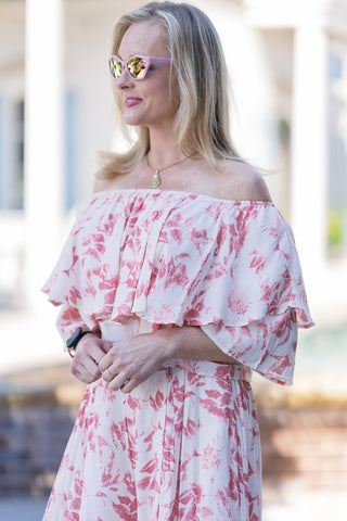 An upper body close-up of a woman wearing the Bellini Brunch Set, highlighting the dreamy elasticized off-the-shoulder neckline that beautifully showcases the collarbones. The top features dramatic, double-layered cascading tiered ruffles in a warm terracotta and peach botanical floral print over a creamy ivory canvas. Styled with pink cat-eye sunglasses and a gold pendant necklace, the image perfectly captures the set's lightweight, crinkle slub texture and carefree coastal Italian aesthetic.