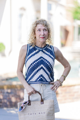 A portrait view focusing on the artisanal texture of the Retro Wave Cropped Knit Top. The image perfectly showcases the heavy-gauge crochet weave and the striking navy and cream chevron stripes. The sleeveless top features a classic crew neckline with a contrast ribbed trim. The woman poses with one hand on her hip, styling the retro top with cream drawstring shorts, stacked gold bracelets, and a woven straw tote bag carrying patriotic sunglasses.