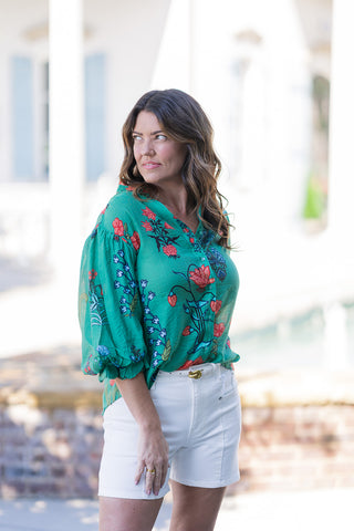 A portrait view of the Jade Botanical Blouse highlighting the flattering split band-collar v-neckline and functional dyed-to-match button-down front. The image showcases the dramatic 3/4-length balloon sleeves finished with gathered, elasticized ruffle cuffs. The premium, lightweight woven fabric displays an intricate, block-print floral motif against a rich jade green base, offering a polished yet artistic flair.