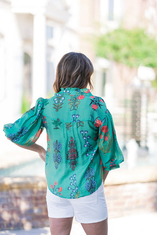 Back view of the Jade Botanical Blouse showcasing the relaxed, effortless drape that skims gently over the figure without clinging. The image highlights the beautiful continuation of the vibrant block-print botanical pattern across the back and the playful volume of the 3/4-length balloon sleeves. Styled with white denim shorts for a comfortable, bright summer aesthetic.