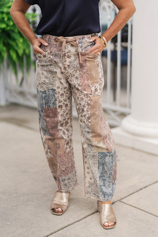 A close-up mid-shot focusing on the design details of the Risen Mocha Mosaic Barrel Pants from the waist down. The image highlights the intricate geometric seams connecting the floral, paisley, and mosaic-printed earth-toned panels. The flexible waistband is equipped with an integrated brown drawstring cord tied at the center, alongside functional front utility patch pockets where the woman has placed her hands.