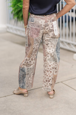 A back view from the waist down of a woman wearing the Risen Mocha Mosaic Barrel Pants, emphasizing the slouchy taper of the barrel silhouette from behind. The continuous mosaic patchwork layout features large patterned panels that sweep across the rear legs to the cropped ankle hemline. The design is finished with classic back patch pockets, a paper brand tag near the waistband, and is styled with gold slide sandals.