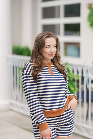 A close-up portrait view of the Yacht Club Lounge Top highlighting the maritime-inspired design elements. The knit sweatshirt fabric showcases a clean navy blue and cream horizontal stripe motif. The functional Henley collar is trimmed with a contrasting warm tan ribbed knit fabric and features small matching buttons. The woman is posed looking to the side with one hand in her pocket, showcasing the solid tan banded waist and cuffs.