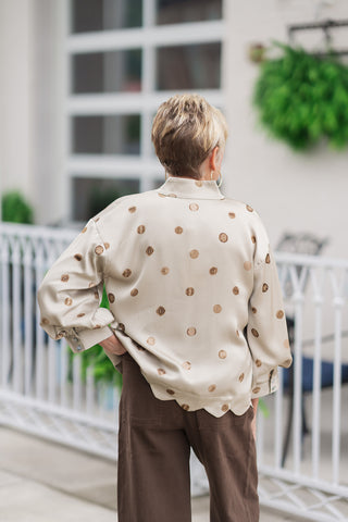 Back view from the waist up of a woman wearing the Sweet Wave Collared Blouse, showcasing the continuous distribution of the textured brown dot print across the back and shoulders. This angle highlights the fluid drape of the relaxed drop-shoulder tailoring, the structure of the standing collar from behind, and the clean finish of the scalloped hem resting against the waistband of her dark brown trousers.