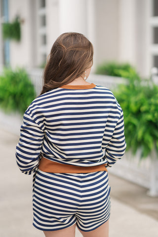 Back view from the waist up of a woman wearing the Yacht Club Lounge Top, showcasing the continuous navy blue and cream maritime horizontal stripe pattern across the shoulders and long sleeves. This angle highlights the comfortable, relaxed fit of the pullover and features a contrasting warm tan ribbed collar and solid tan banded hemline that separates the top from the matching lounge shorts.