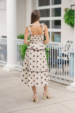 A back view of a woman standing on an outdoor patio wearing the Espresso Truffle Polka Dot Dress, showcasing the dress's functional and structural design from behind. The upper back bodice features a comfortable, smocked elastic panel to ensure a secure fit, bordered by dark brown contrast piping. The cream and espresso-brown polka dot pattern flows continuously through the peplum waist and down to the tiered ruffled midi hemline, styled with chunky platform heels.