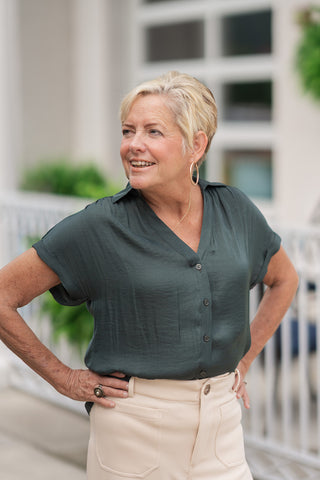An upper-body portrait view of the Mossy Morning Collared Blouse showcasing the premium fabric's texturized sheen. The design features a classic pointed shirt collar that frames a plunging V-neckline, secured by tonal buttons down the front placket. The short, cuffed cap sleeves are finished with a fixed rolled edge. The woman poses with her hands on her hips, accessorized with gold hoop earrings and a delicate necklace.