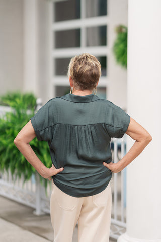 Back view from the waist up of a woman wearing the Mossy Morning Collared Blouse, highlighting the fluid drape of the relaxed silhouette. The deep hunter green satin-blend fabric features a gathered back yoke that introduces soft pleating across the shoulders. The top is worn untucked to display the curved, straight hemline as it sits smoothly against the high-waisted band of her cream trousers.