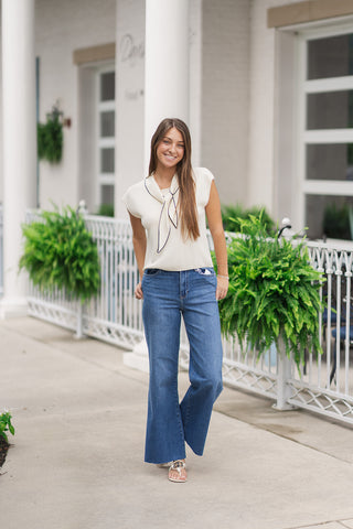 A full-length view of a smiling woman walking forward in the cream French Riviera Sleeveless Sweater. The top is styled for a casual, European-inspired look with wide-leg blue denim jeans and white flat thong sandals. The clean hemline rests neatly at the hips, emphasizing a chic and relaxed resort-ready aesthetic.