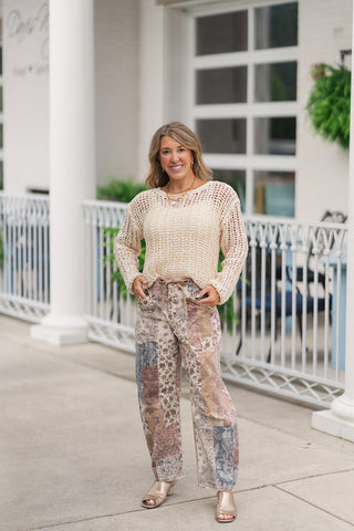 A full-length front view of a smiling woman standing on an outdoor patio wearing the Dear John Light Beige Adela sweater. The premium pullover features a breathable, open-weave crochet style grid pattern in a warm, neutral light beige tone with a wide boat neckline and long sleeves. It is styled for a relaxed, bohemian boutique look over earth-toned mosaic patchwork wide-leg trousers, metallic gold block-heel slide sandals, and a layered gold charm necklace.