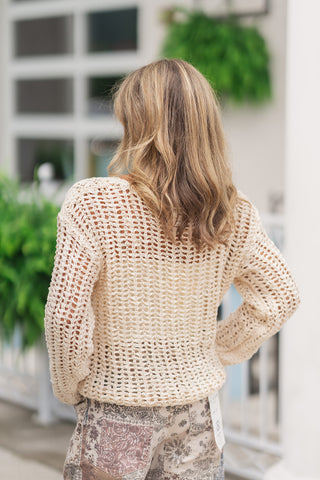 Back view from the waist up of a woman wearing the Dear John Light Beige Adela sweater, highlighting the clean grid symmetry of the open-knit pattern across the reverse side. The slightly cropped hemline rests naturally against the high-rise waistband of her floral mosaic patchwork pants, showing off the relaxed, slouchy shape and continuous open-weave design from behind.