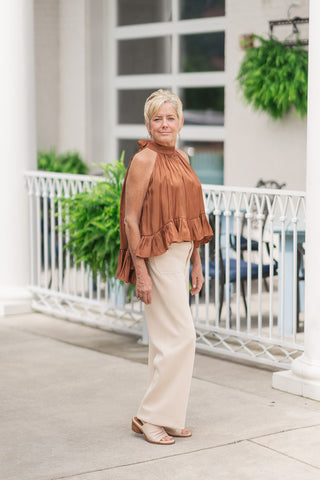 A full-length side view of a woman standing on an outdoor patio wearing the Toasted Cinnamon Shimmer Top. The top is rendered in a warm toasted-cinnamon orange-brown hue with an iridescent finish. Cut in a dramatic high-low silhouette, the sleeveless top sits shorter in the front and lengths into a flowing, tiered ruffle back hem. The outfit is styled for a polished resort aesthetic with cream high-waisted wide-leg trousers and neutral open-toe block heel sandals.