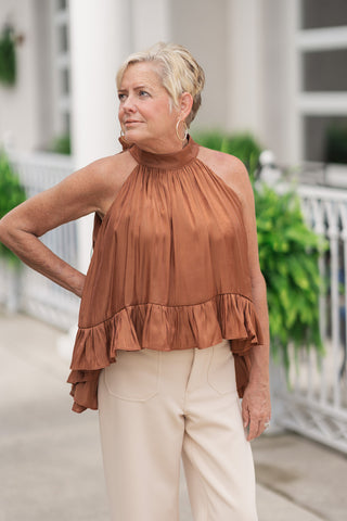 An upper-body portrait view of the Toasted Cinnamon Shimmer Top, showcasing the luminous sheen of the premium metallic-effect fabric. The top highlights a high halter neckline accented by elegant gathers that flow into a fluid, breezy silhouette. The ruffle hem is visible at the front waist, styled untucked over light beige trousers and paired with large gold hoop earrings.