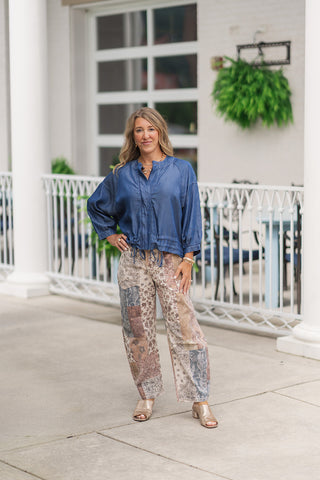 A full-length front view of a smiling woman posing on an outdoor patio wearing the Seaside Stroll Bomber Jacket. This lightweight, unlined jacket is made from a dark denim blue Tencel fabric with a soft, subtle sheen. It features a high stand collar, drop-shoulder $3/4$ sleeves, and a gathered elastic waistband with an adjustable drawstring hem. The jacket is styled zipped up and cinched with earth-toned mosaic patchwork barrel pants, gold block-heel slide sandals, and a gold link necklace.