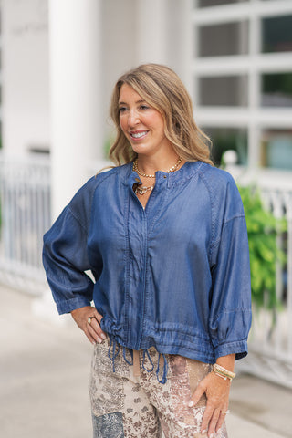 A portrait-style mid-shot of the Seaside Stroll Bomber Jacket, highlighting the fluid, premium drape of the dark denim blue Tencel fabric. The view showcases the minimalist stand collar, a partial front zipper closure, and loose-fitting drop-shoulder sleeves that taper into structured, gathered cuffs. The adjustable drawstring is cinched at the front waist, styled with layered gold coin and link chain necklaces.