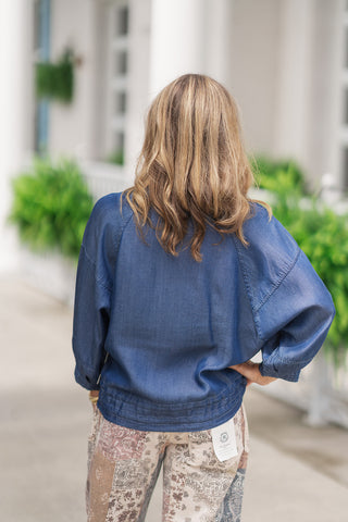 Back view from the waist up of a woman wearing the Seaside Stroll Bomber Jacket, emphasizing the relaxed fit and blouson-style bubble effect of the design. This angle highlights the continuous, clean look of the dark blue Tencel fabric across the shoulders and the wide, elastic-gathered bottom hemline. The jacket is styled untucked to showcase how the gathered hem rests smoothly against the waistband of her patterned patchwork trousers.