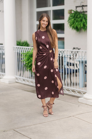 A full-length view of a smiling woman posing on an outdoor patio in the Milano Metallic Twist Midi Dress. The dress features a classic polka dot print with large light pink dots set against a rich chocolate brown woven fabric. The sleeveless silhouette showcases an artfully draped front with an asymmetrical wrap effect, beautifully anchored at the waist by a polished gold-tone metal hardware accent. The midi-length skirt features an elegant side-front hem slit, styled with brown strappy block-heeled sandals