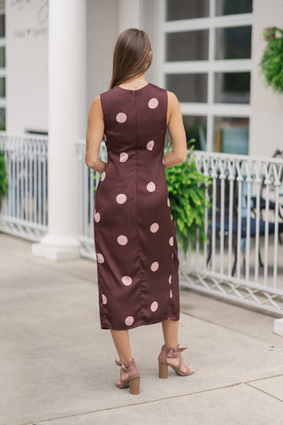 Back view of a woman standing on an outdoor patio wearing the Milano Metallic Twist Midi Dress, showcasing the clean, uninterrupted silhouette of the dress. The large light pink polka dots are evenly distributed across the chocolate brown fabric from the modest round neckline down to the straight midi-length hem. The view highlights the tailored, non-sheer fit, a hidden central back zipper closure, and the back tie details of her brown heeled sandals.