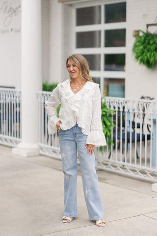 A full-length front view of a smiling woman standing on an outdoor patio wearing the Dear John Starboard Taylor jeans. These straight-leg jeans feature a light-to-medium blue denim wash accented by delicately distressed, light-wash star patch appliques scattered across the front of the legs. The whimsical look is styled with a white long-sleeve blouse featuring cascading front ruffles and flared trumpet cuffs, cream-colored flat slide sandals, and a gold link charm necklace.