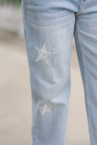 A detailed macro close-up focusing on the artisanal embroidery of the Dear John Starboard Taylor jeans. The image highlights a singular, light-wash denim star applique securely stitched onto the front thigh section, showcasing the rich texture of the embroidery thread and the surrounding subtle denim whiskering.