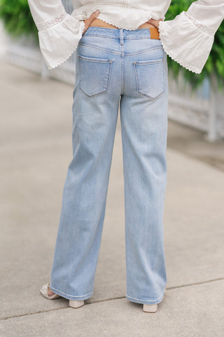 Back view from the waist down of a woman wearing the Dear John Starboard Taylor jeans, showcasing the flattering straight-leg fit and clean rear pocket alignment from behind. The light-to-medium wash stretch denim features classic back patch pockets and a small tan leather brand patch on the right side of the waistband. The model poses with her hands on her hips, highlighting the cropped ankle-length hemline styled with cream sandals.