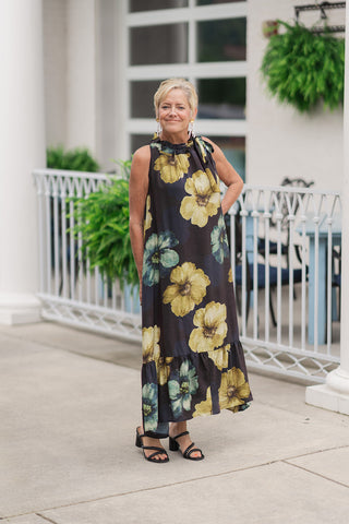 A full-length front view of a smiling woman standing on an outdoor patio wearing the Golden Moss Halter Midi Dress. This sleeveless floral midi dress showcases a large-scale, painterly floral pattern featuring shades of chartreuse-yellow, cream, and deep sage-plum against a dark grounding background. The design features an elegant high halter neckline with a soft ruffle trim and a fluid silhouette that flows into a tiered ruffle hemline. The look is styled for a sophisticated warm-weather event with strappy