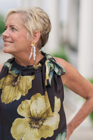 A portrait-style side profile mid-shot highlighting the upper structural features of the Golden Moss Halter Midi Dress. The lightweight, breathable fabric highlights a high halter neck gathered into a charming ruffle trim. A thick customizable self-tie fabric strap loops into a bow at the side of the neck. The woman is looking off-camera, wearing long, cascading white baroque pearl drop earrings suspended from gold floral studs.