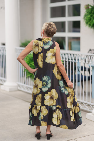 Back view of a woman standing on an outdoor patio wearing the Golden Moss Halter Midi Dress, highlighting the continuous print alignment and graceful movement of the garment. The large-scale chartreuse floral design runs seamlessly from the high ruffled back neck down to the tiered ruffled midi hemline. The woman has one hand placed casually on her hip, showcasing the airy drape and non-restrictive, sleeveless shift silhouette.