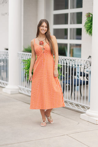 A full-length view of a woman smiling and posing on an outdoor patio wearing the Summer Ripple Midi Dress, a vibrant, sleeveless plaid midi dress. The design features a lively orange and pink gingham check pattern, a structured under-bust seam, and a keyhole cutout at the neckline secured with a delicate tie closure. The look is styled for a breezy summer aesthetic with white flat slide sandals and an oversized statement coral-motif pendant necklace in front of a white columned storefront.