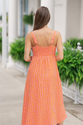 Back view from the waist up of a woman wearing the Summer Ripple Midi Dress, showcasing the comfortable smocked elastic back panel that ensures a perfect fit. The view highlights the continuous orange and pink gingham plaid pattern, matching adjustable shoulder straps with gold-toned hardware, and a clean, straight back neckline above a gently flowing, tiered midi skirt.