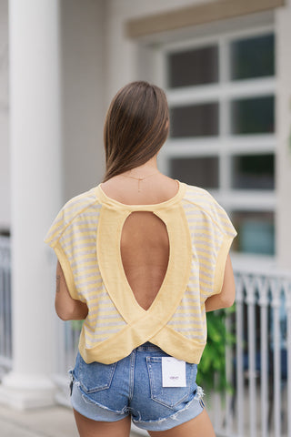Back view from the waist up of a woman wearing the Lemon Drop Lounger Top, highlighting the dramatic open-back crossover design. The vertical oval-shaped cutout exposes the bare back, framed by the yellow striped knit fabric overlapping at the bottom hem. The shirt is paired with high-waisted distressed denim shorts featuring a white brand tag on the back right pocket.