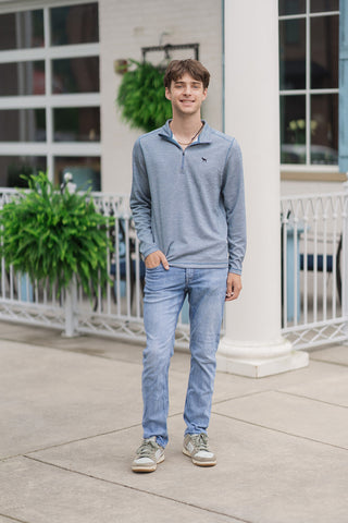 A full-length front view of a smiling young man standing outdoors on a concrete patio wearing the Slate Harbor Zip Pullover. The long-sleeve pullover is featured in a textured blue and grey melange weave, styled with light-wash slim-fit denim jeans and olive-green and white sneakers in front of a white columned entryway.